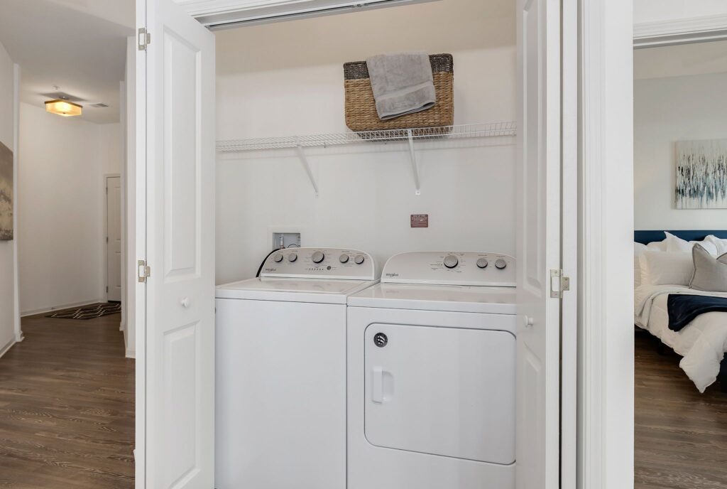 Laundry area with a washer and dryer, a shelf above holding a basket and folded towels. White bi-fold doors open to hardwood flooring and a glimpse of a bedroom on the right.