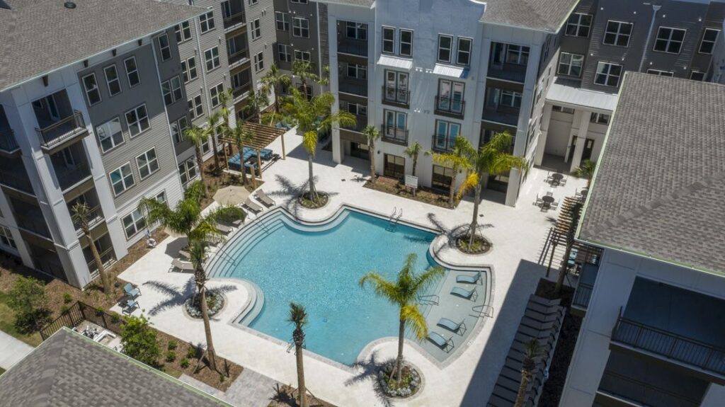 Aerial view of a modern apartment complex with a central outdoor swimming pool, surrounded by palm trees and lounge chairs.