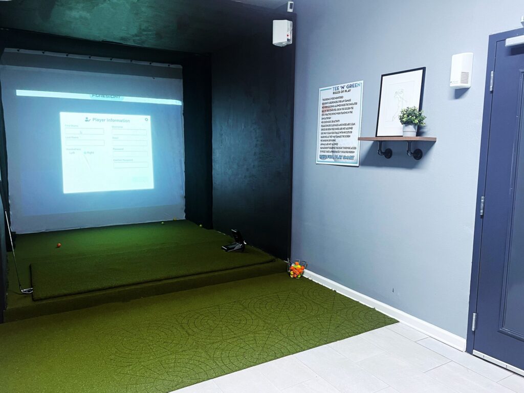 Indoor golf simulator setup with a screen displaying player information, green turf on the floor, a basket of balls, and wall decor.