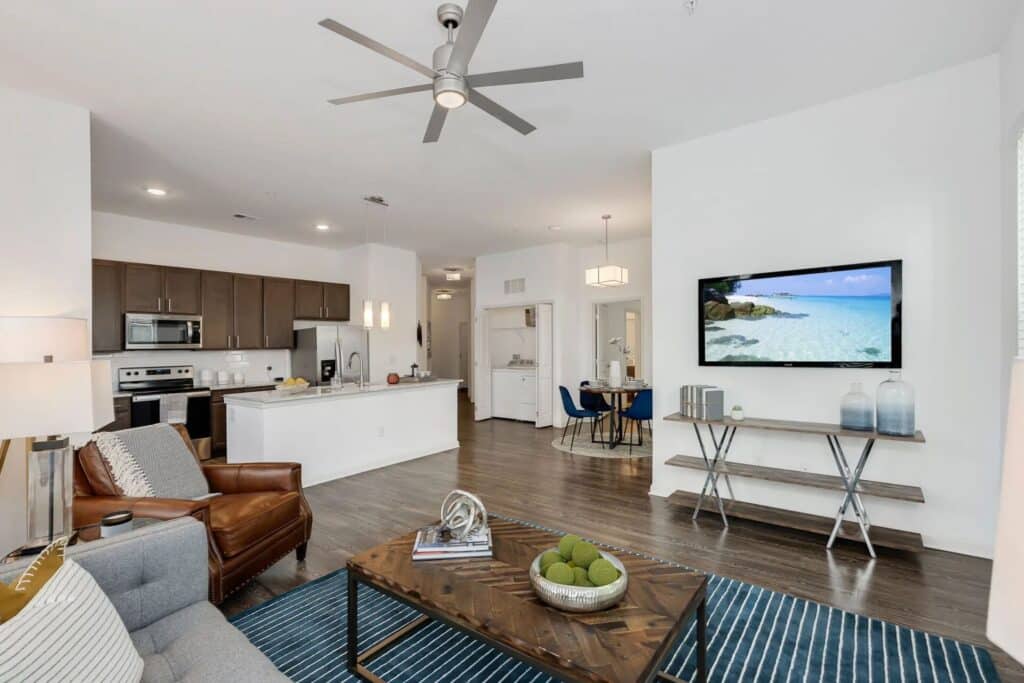 Modern living room with a ceiling fan, TV, and light decor. Open kitchen with dark cabinets and stainless steel appliances in the background. Blue rug on wooden floor.