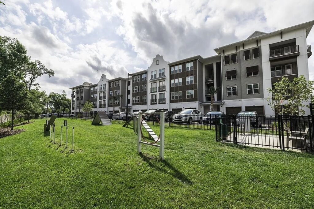 A fenced dog park with agility equipment is in front of a large, multi-story apartment complex. The sky is partly cloudy.