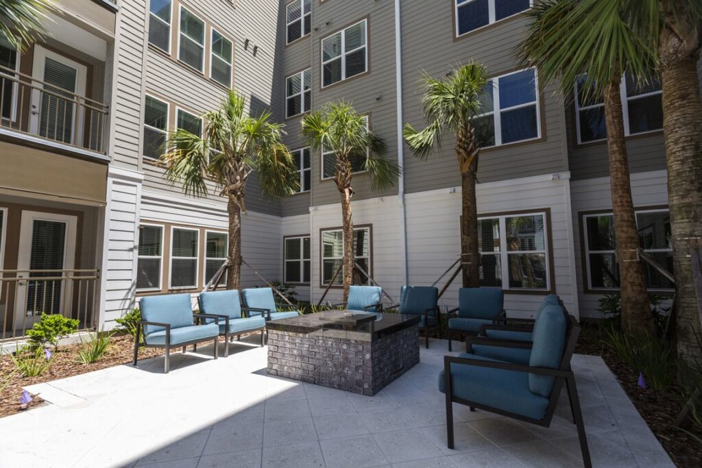 Courtyard with palm trees, blue cushioned chairs, and a central fire pit, surrounded by multi-story gray and white buildings.