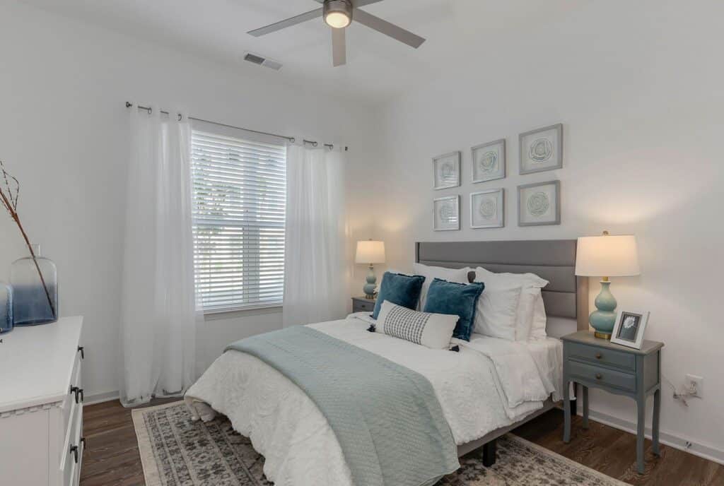 A bedroom with a neatly made bed featuring blue and white bedding, flanked by two lamps on side tables. A window with sheer curtains is on the left, and four framed pictures are on the wall.