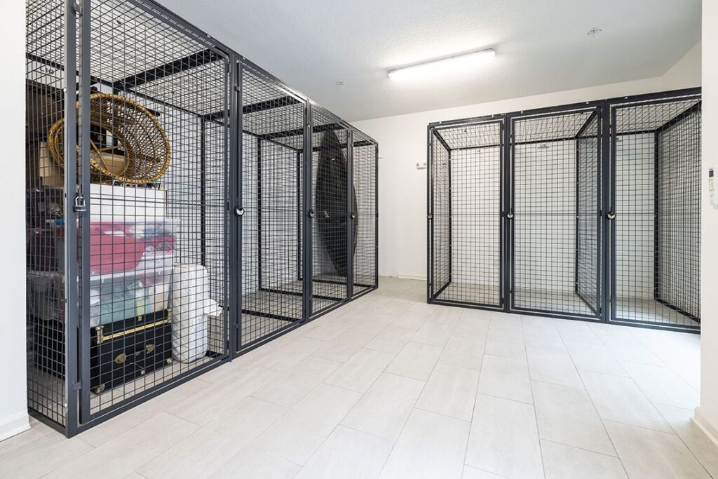 Storage room with wire mesh cabinets, filled with assorted items such as boxes and a hula hoop. The room has a tiled floor and fluorescent lighting.