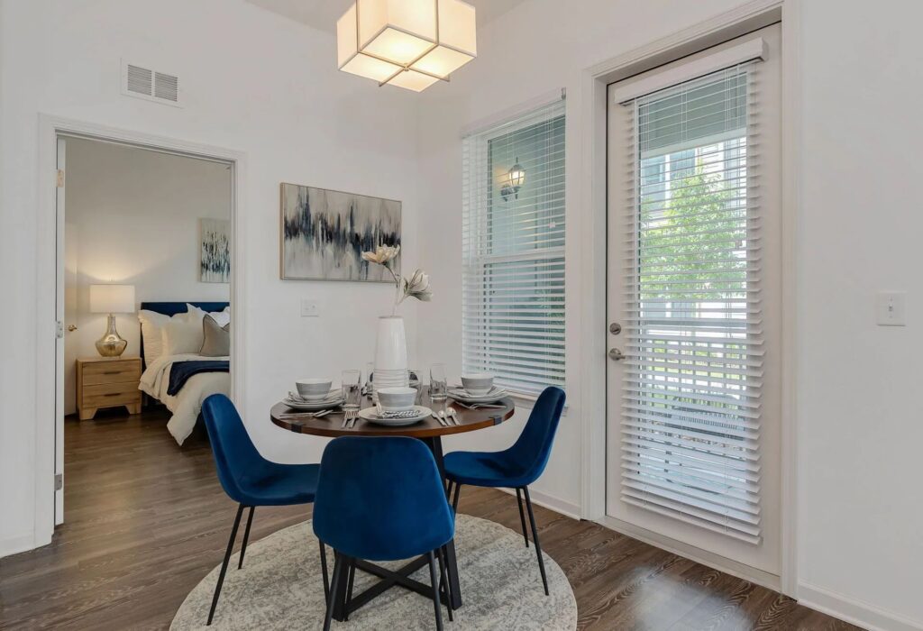 Dining area with a round table set for four, surrounded by blue chairs on a circular rug. A bedroom is visible through an open door, and large windows with blinds are on the right.