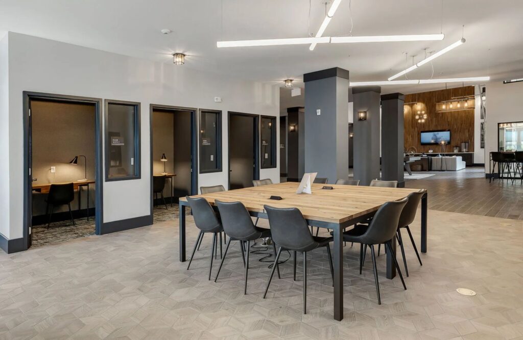 Modern office space with a large wooden table surrounded by chairs in the center, individual work booths on the left, and a lounge area with a TV in the background.