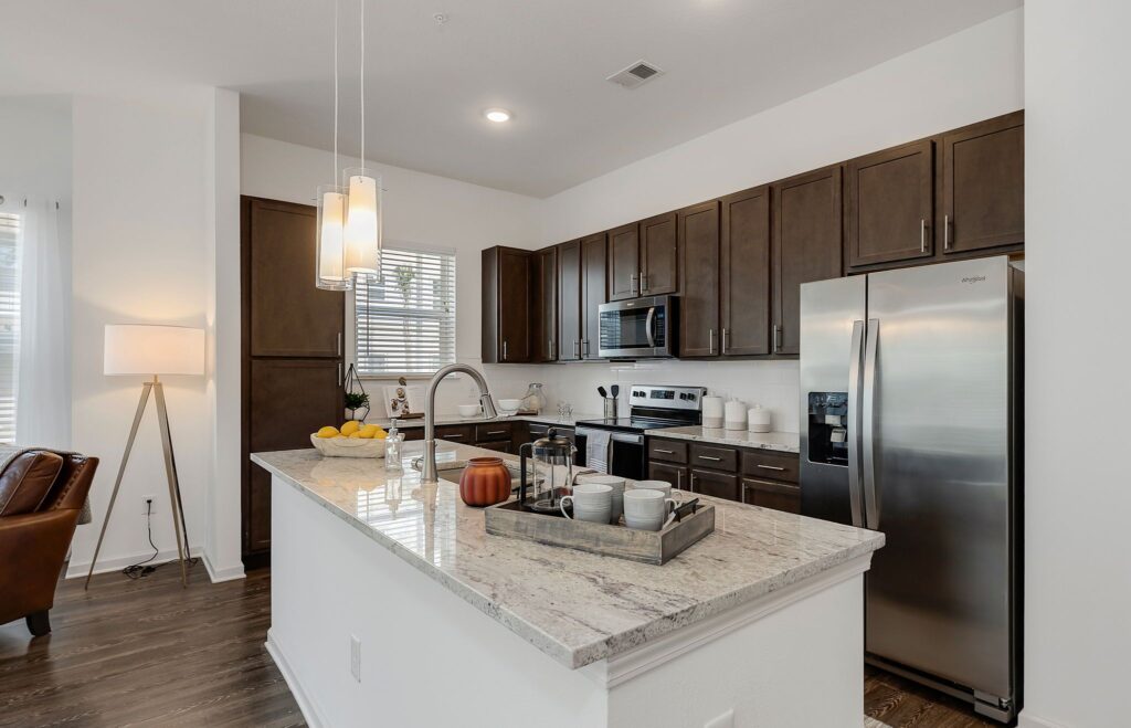 Modern kitchen with dark wood cabinets, stainless steel appliances, and a marble countertop island. Pendant lights hang above the island, which has a tray with cups and a decorative jar.