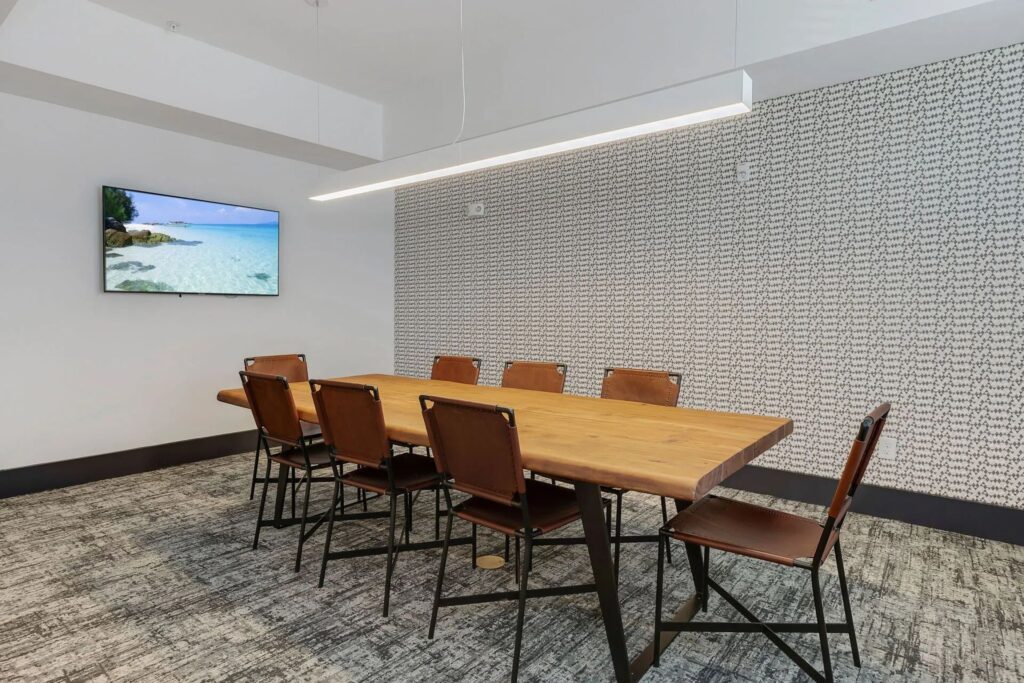 A modern conference room with a wooden table, eight brown chairs, a wall-mounted screen displaying a beach scene, and patterned wallpaper.