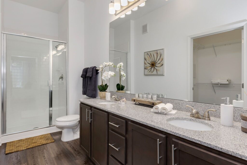 Modern bathroom with dual sinks, granite countertop, a large mirror, shower with glass door, and decor including orchids and towels.