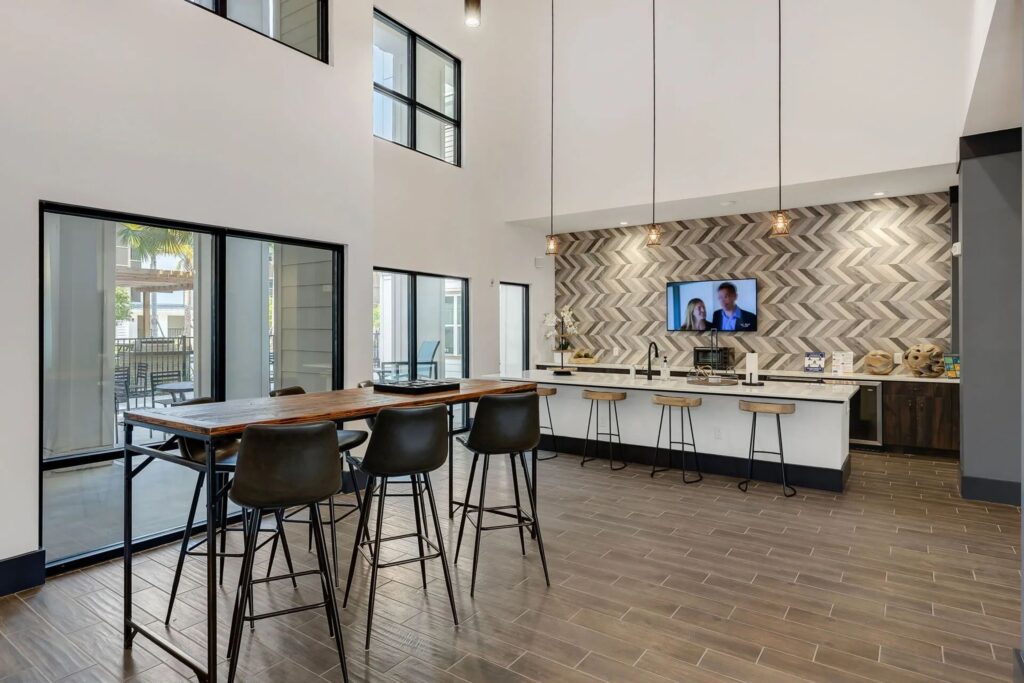 Spacious modern kitchen with high ceilings, island seating, pendant lights, and a herringbone tile backsplash, featuring large windows and a wall-mounted TV.