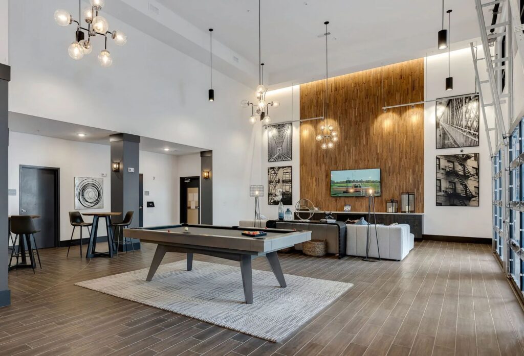 A modern lounge with high ceilings, a pool table on a rug, a seating area with a TV, and wall art. Hanging lights illuminate the space.